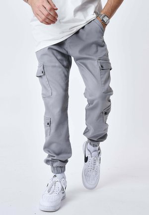 Personne portant un pantalon jogger cargo gris, des baskets blanches avec un logo noir, des chaussettes blanches et une chemise blanche, une main dans la poche.