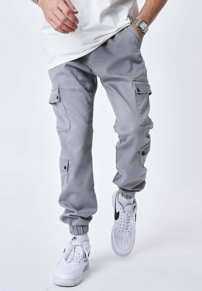 Personne portant un pantalon jogger cargo gris, des baskets blanches avec un logo noir, des chaussettes blanches et une chemise blanche, une main dans la poche.