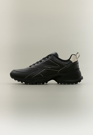 Baskets en cuir noir avec semelle épaisse, languette de talon blanche et logo Calvin Klein sur le côté, présentées de profil sur fond neutre.