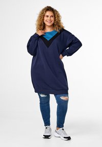Vestito felpa oversize color navy con accento a forma di V nera e pannello teal; abbinato a jeans blu distrutti e sneaker grigie.