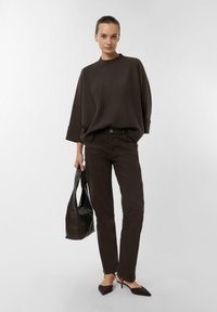 Brązowy oversize'owy sweter z wysokim kołnierzem, zestawiony z dopasowanymi brązowymi jeansami. Modelka niesie czarną skórzaną torbę typu tote i nosi czarne spiczaste buty.