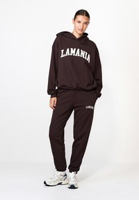 Hnedý súprava mikiny a teplákov s bielym logom "LAMANIA". Materiál vyzerá mäkko; uvoľnený strih s rebrovanými manžetami a lemom. Na sebe sú športové topánky.