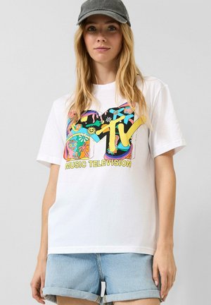 Jeune femme portant une casquette grise, un t-shirt blanc MTV Music Television avec un motif coloré, et un short en jean bleu clair retroussé.