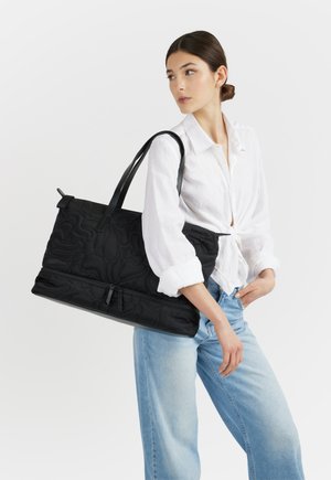 Donna con camicetta bianca e jeans blu che porta una grande borsa tote nera con texture trapuntata sulla spalla, su sfondo bianco.