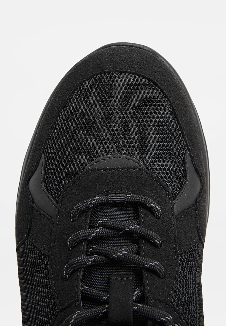 Schwarzer Sport-Schuh mit einem Obermaterial aus Mesh, Suede-Akzenten und einer gepolsterten Sohle. Auch die Schnürsenkel und die verstärkte Zehenkappe sind sichtbar.