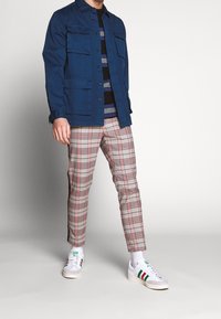 Blå canvasjacka med flera fickor, randig skjorta, grå rutiga tapered byxor och vita sneakers med gröna, röda och svarta accentfärger.