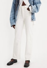 Jeans blancos de cintura alta con un ajuste relajado y bajos deshilachados, combinados con una chaqueta de mezclilla azul y mocasines negros.