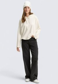 Világos krémszínű pulóver kapucnival és széles ujjakkal, fekete bő szárú nadrággal és fehér tornacipővel párosítva, fehér kötött sapkával kiegészítve.