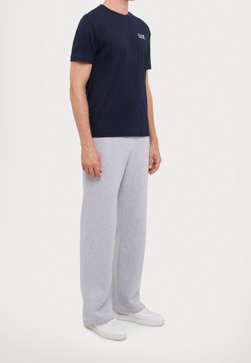 Man som bär en marinblå EA7-t-shirt, ljusgrå vida sweatpants och vita sneakers, stående framför en enfärgad bakgrund.