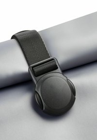 Cinturino in nylon nero con una fibbia rotonda e testurizzata che fissa una superficie grigia opaca e liscia, probabilmente un oggetto o materiale arrotolato.