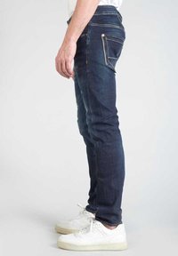 Jeans de denim ajustados en un oscuro tono azul. Incluyen bolsillos frontales en ángulo y un bolsillo trasero con cremallera con herrajes plateados.