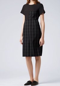 Černé šaty s přiléhavým vrškem a sukní v kostkovaném vzoru. Hladká tkanina, krátké rukávy a délka po kolena. Ploché černé boty.