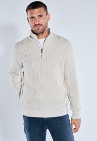 Beige Zip-Up-Pullover mit geripptem Struktur, langen Ärmeln und hohem Kragen, kombiniert mit dunkelblauen Jeans. Abgerundet mit minimalen Akzenten.
