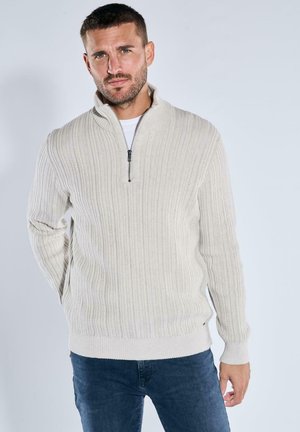 Beige Zip-Up-Pullover mit geripptem Struktur, langen Ärmeln und hohem Kragen, kombiniert mit dunkelblauen Jeans. Abgerundet mit minimalen Akzenten.