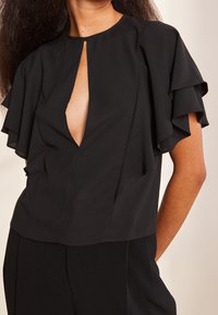 Donna che indossa una blusa nera con maniche corte a strati e scollatura a chiave frontale, abbinata a pantaloni neri.