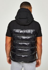 Mężczyzna w czarnej kamizelce bez rękawów z kapturem, widziany z tyłu, w połączeniu z jasnoszarymi spodniami na neutralnym tle.