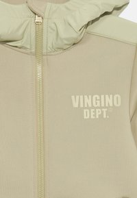 Beige zip-up hoodie with a high collar, featuring a smooth texture and the logo "VINGINO DEPT." in a contrasting cream color. Zipper details visible.