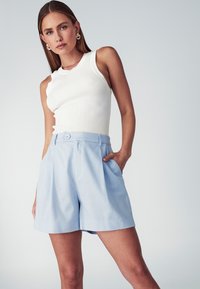 Bílý žebrovaný top s kulatým výstřihem, který je zkombinován s světle modrými skládacími šortkami, jež mají boční kapsy a detail s knoflíkem na pase.