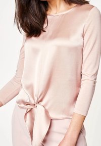 Ljust rosa satinblus med rund halsringning, långa ärmar och en knuten detalj vid midjan. Slät textur och glänsande ytfinish.