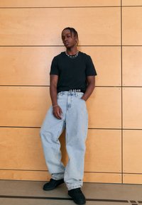 Zwart T-shirt, lichtblauwe wijde spijkerbroek met een zichtbaar logo op de tailleband en zwarte schoenen. Staand tegen een houten paneelachtergrond.