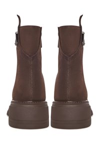 Paio di stivaletti in suede marrone con linguette e fibbie, visti da dietro, su suole in gomma spessa contro uno sfondo bianco.