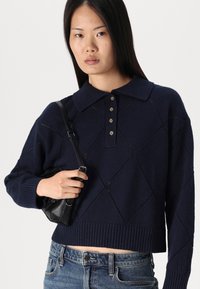 Mornariće plavi pleteni pulover s ovratnikom, dijamantnim uzorkom i rebrastim rubom. Model drži malu crnu torbu i nosi traperice.