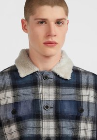 Young man with short hair wearing a blue and white plaid jacket with a white sherpa collar, buttoned up at the front.