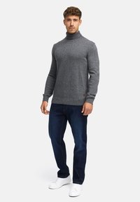 Pull col roulé gris avec col et poignets côtelés, assorti à un jean en denim foncé et des baskets blanches. Le mannequin se tient devant un fond uni.