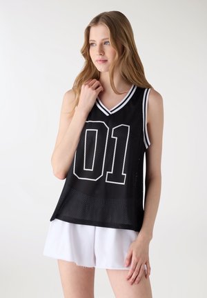 Giovane donna che indossa una maglia nera senza maniche in rete con il numero "01" bianco e pantaloncini bianchi, in piedi davanti a uno sfondo chiaro uniforme.