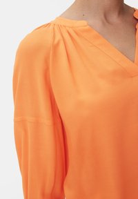 Orange Bluse mit V-Ausschnitt, gerafften Schulternähten und einer weichen, glatten Textur. Lange Ärmel mit dezenten Manschetten-Details.