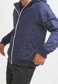 Giacca blu navy con zip e cappuccio, caratterizzata da una cerniera bianca, due tasche frontali e polsini a costine. Materiale leggero e lucido.