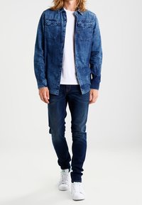 Blårutigt denimjacka över en vit t-shirt, kombinerad med mörka jeans och vita sneakers, med två bröstfickor och knapppdetaljer.