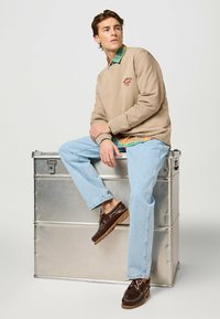 Felpa beige con logo, sovrapposta a una camicia a scacchi verde e arancione, abbinata a jeans azzurri chiari e scarpe da barca marroni. Seduto su una scatola di metallo.