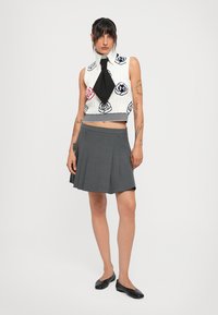 Kvinne med mørkt hår og tatoveringer har på seg en hvit ermeløs topp med blomstermønster og et svart slips, grå folderett skjørt, og svarte flate sko.