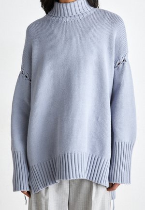 Kvinde iført en oversized lyseblå ribstrikket polotrøje med udskæringsdetaljer på ærmerne og lysegrå bukser med nålestriber.
