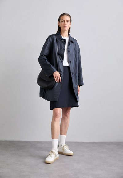 Navy wasserfeste Jacke mit Knöpfen, kombiniert mit einem weißen T-Shirt und einem navyfarbenen Rock. Weiße Sneaker und eine schwarze Umhängetasche runden den Look ab.
