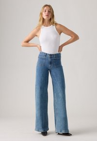 Hoge taille, wijde jeans in lichtblauwe denim met vier zakken; gedragen met een witte mouwloze top en zwarte hakken.
