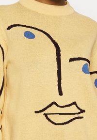 Gul stickad tröja med en abstrakt svart linje ansiktsdesign som har blå ovala ögon och stiliserade läppar på framsidan.