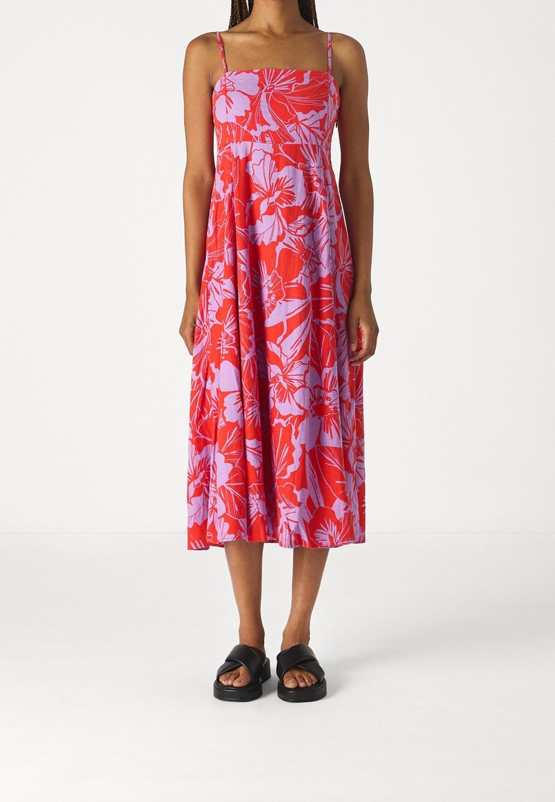 Une femme se tient debout vêtue d'une robe florale rouge vive avec des bretelles fines, associée à des sandales noires, sur un fond neutre.