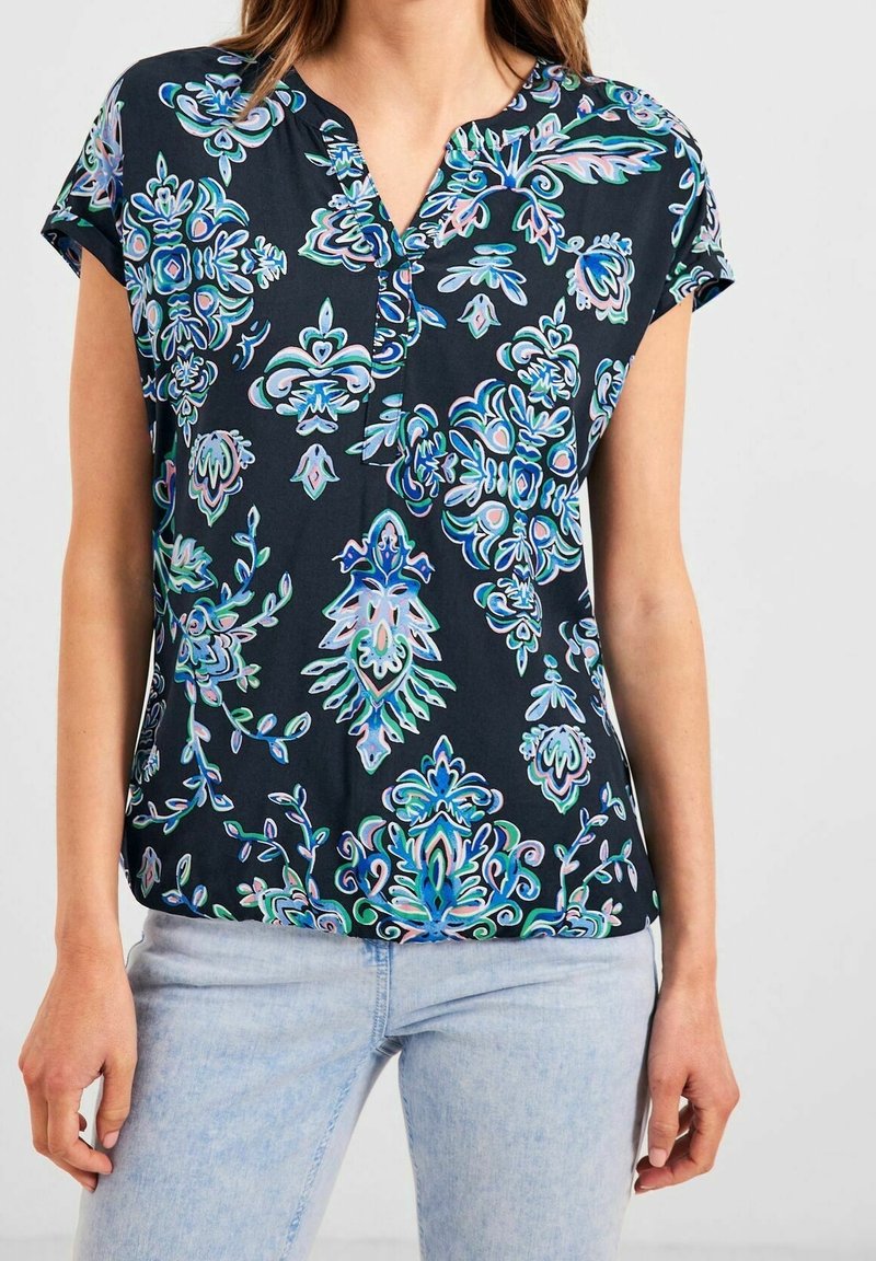 Svart kortärmad blus med livfulla blå och gröna blommönster, en urgröpning vid halsen och en lös passform, matchad med ljusblå jeans.