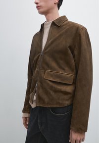 Brun ruskindsjakke med lynlåsfront, spids krave og en lomme. Båret over en beige sweater og parret med sorte jeans.