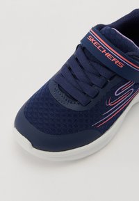 Zapatilla deportiva azul marino con parte superior de malla, cordones, correa de gancho y bucle, y acentos rosas. Presenta una superficie texturizada y una suela acolchada.