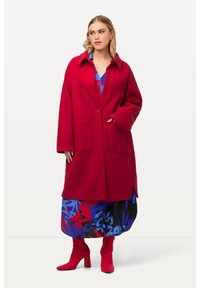 Manteau en laine rouge avec un col, fermeture à un bouton, deux poches avant et fentes sur les côtés, porté sur une robe bleue à fleurs et des bottes rouges.
