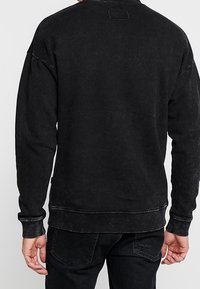 Felpa nera con texture slavata, maniche lunghe, polsini a costine e scollo rotondo. Presenta una piccola toppa quadrata sul retro.