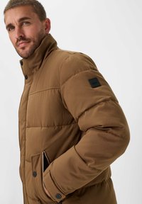 Giacca puffa marrone con colletto alto, tasche con zip e patch logo nero. Il tessuto testurizzato e i dettagli con bottoni arricchiscono il design.