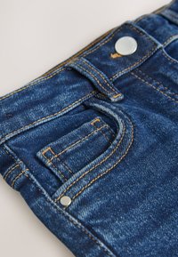 Tmavomodré rifle z denimu s viditeľným prešívaním, malou prednou kapsou a strieborným gombíkom v páse. Hladká textúra s klasickým strihom.