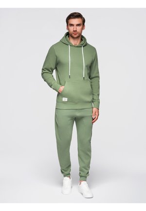 Mann steht, trägt grünen Hoodie und passende Jogginghose mit weißen Sneakers vor einfachem hellem Hintergrund.