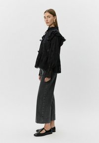 Blusa negra con volantes y detalles recortados, combinada con jeans de cintura alta gris oscuro y zapatos planos negros, sobre un fondo liso.