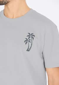 Vaalea harmaa t-paita, jossa on pyöreä pääntie ja vasemmassa rinnassa musta palmu-kuva. Pehmeää puuvillamateriaalia, lyhyet hihat.