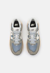 Une paire de baskets New Balance grises, bleues et blanches avec des lacets, vue de dessus sur un fond blanc.